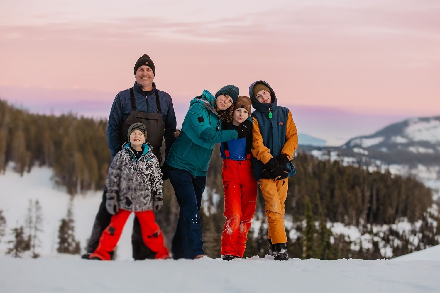 Friends in bright winter gear pose together against snowy mountain backdrop at sunset.