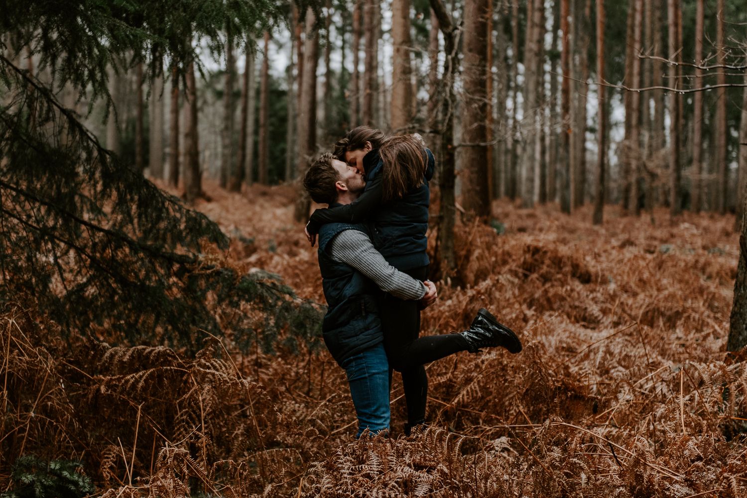 A couple are stood amongst autumnal, golden ferns. The man is lifting the lady up and they are kissing.
