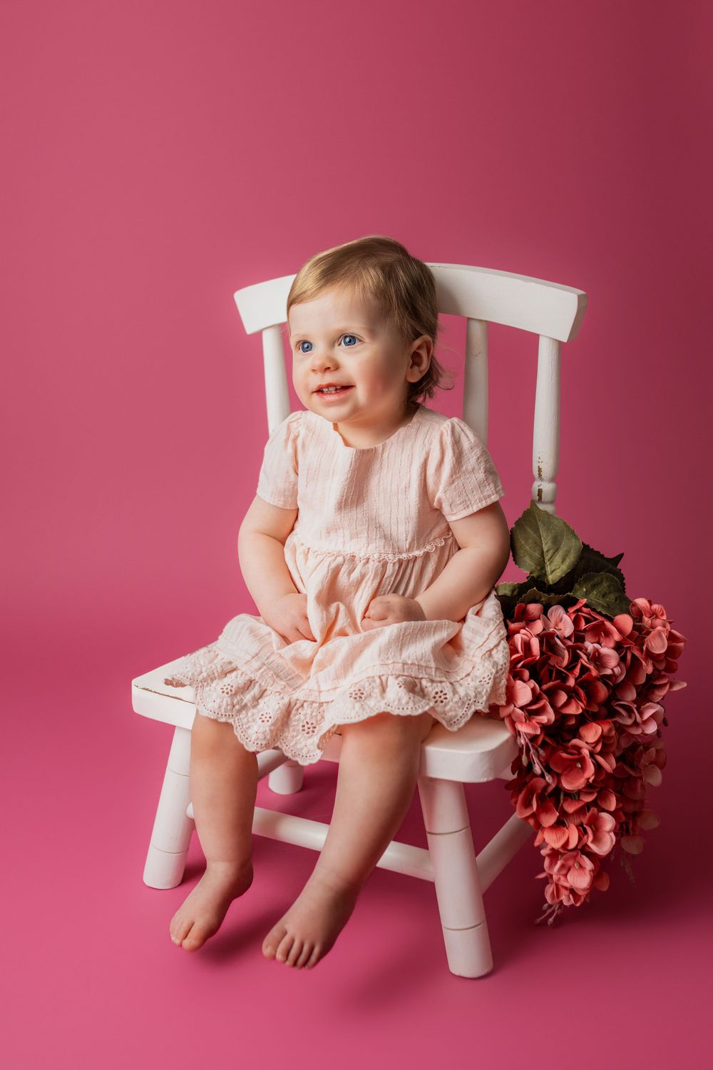 A small child sits on a white wooden chair wearing a pink dress with red flowers beside them on a pink backdrop in cedar park tx baby photos