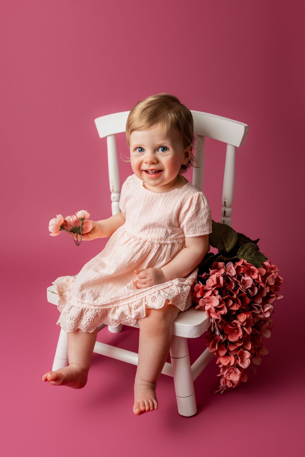 Smiling baby in pink dress sitting on white wooden chair with pink flowers against rose-colored backdrop in cedar park tx baby photo session