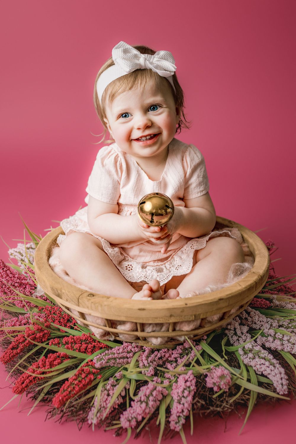 A baby in a pink dress sits in a wooden basket surrounded by pink flowers against a rosy background in cedar park tx baby photoshoot