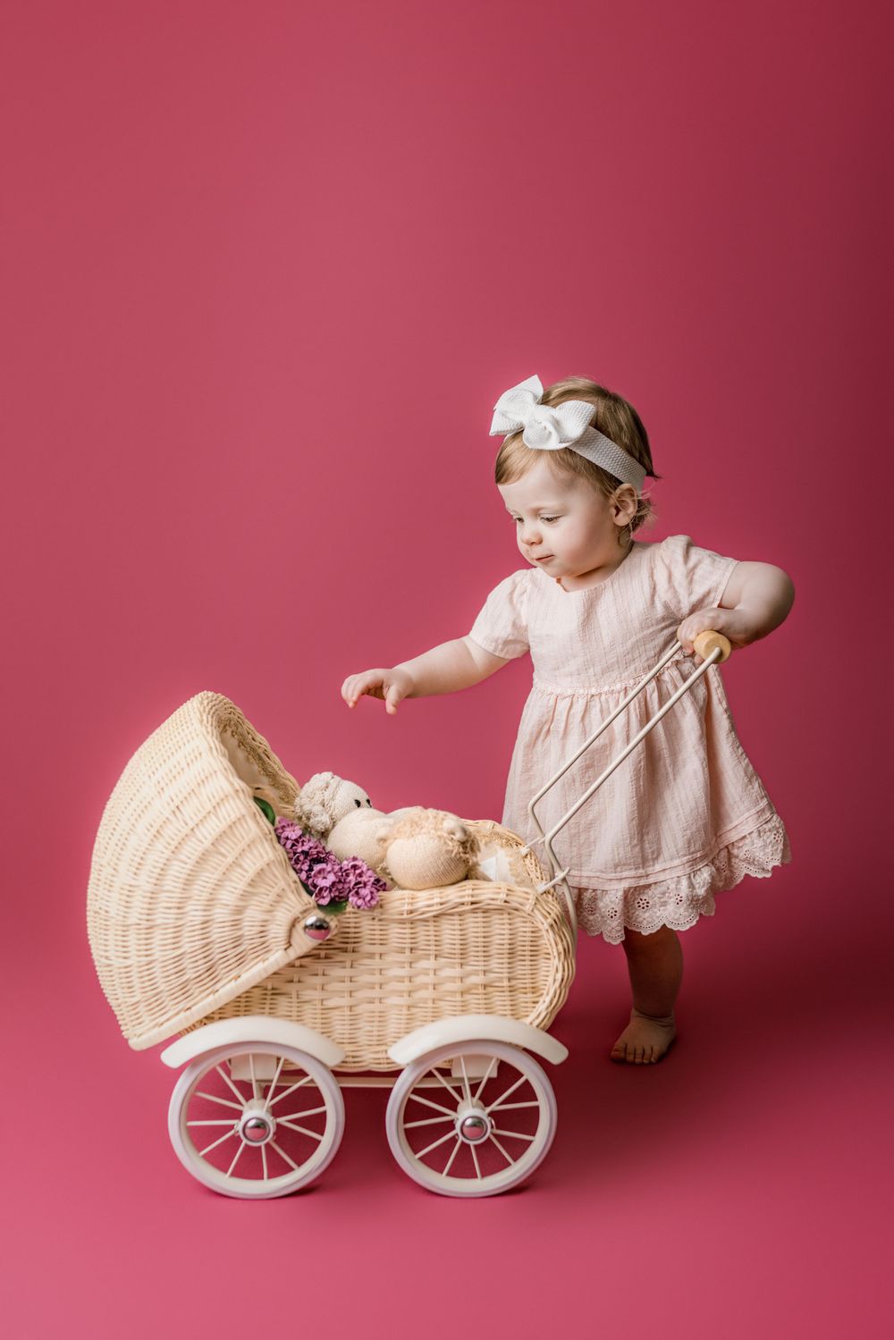 Baby wearing pink lace dress and bow headband plays with vintage wicker doll carriage against pink background in cedar park tx baby photos