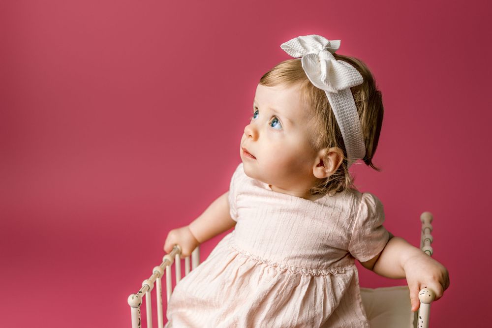 A small child in a pink dress and white bow sits in a white vintage chair against a pink backdrop in cedar park tx first year photo session