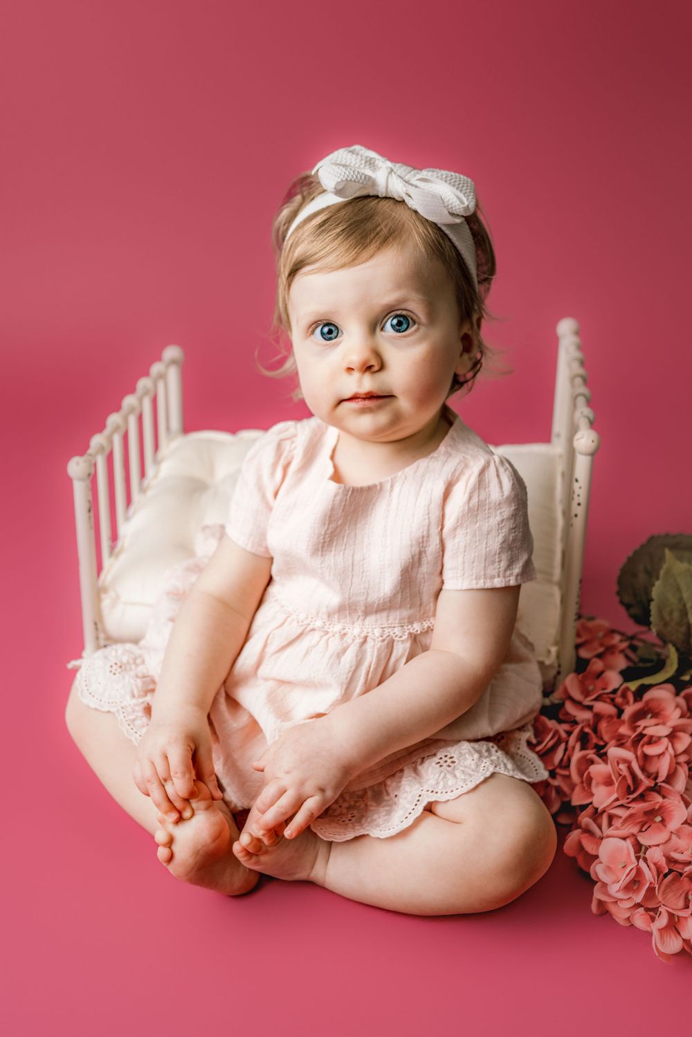 A baby in a pink dress sits on an antique white chair against a pink backdrop with flowers in cedar park tx milestone photo session