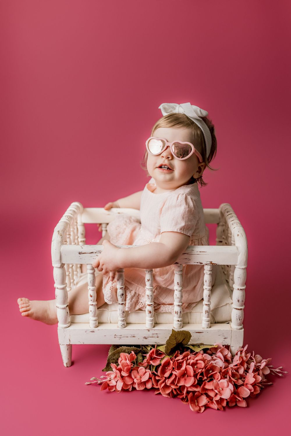Small baby wearing pink dress and sunglasses poses on vintage white bench with pink flowers against rose colored backdrop in cedar park tx