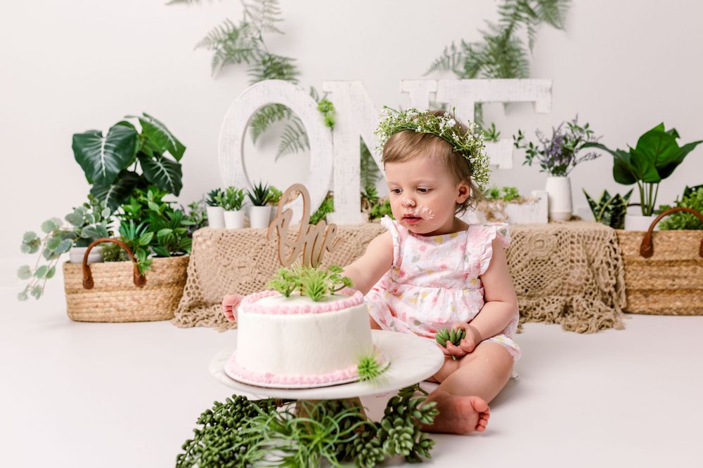 Baby wearing a white dress sits on floor with pink birthday cake surrounded by greenery and potted plants for cake smash photo in cedar park