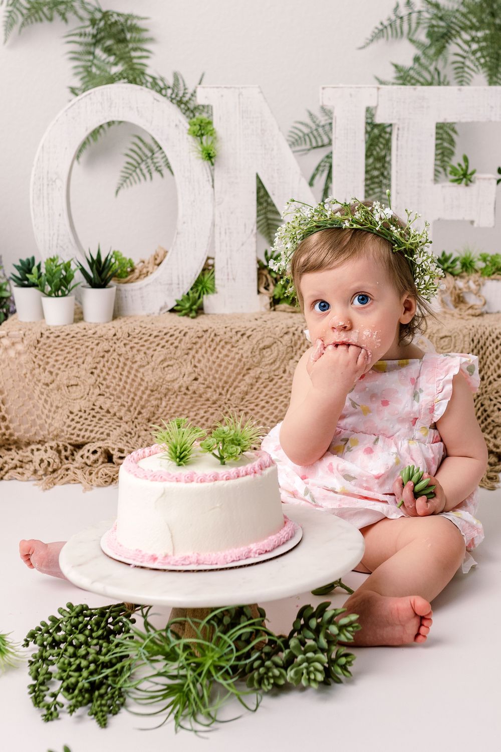 A baby in a floral dress sits next to a white cake with greenery decorations and large ONE letters in the background in cedar park tx