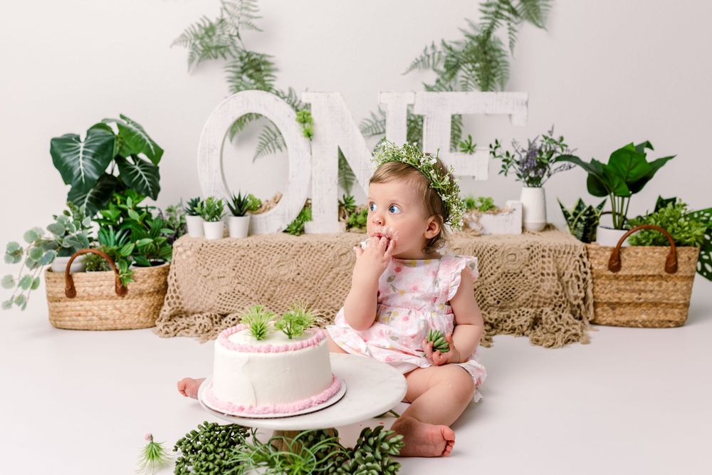 Baby sits on floor at cake smash photoshoot with greenery decor and white letters spelling ONE in background in cedar park tx photo studio