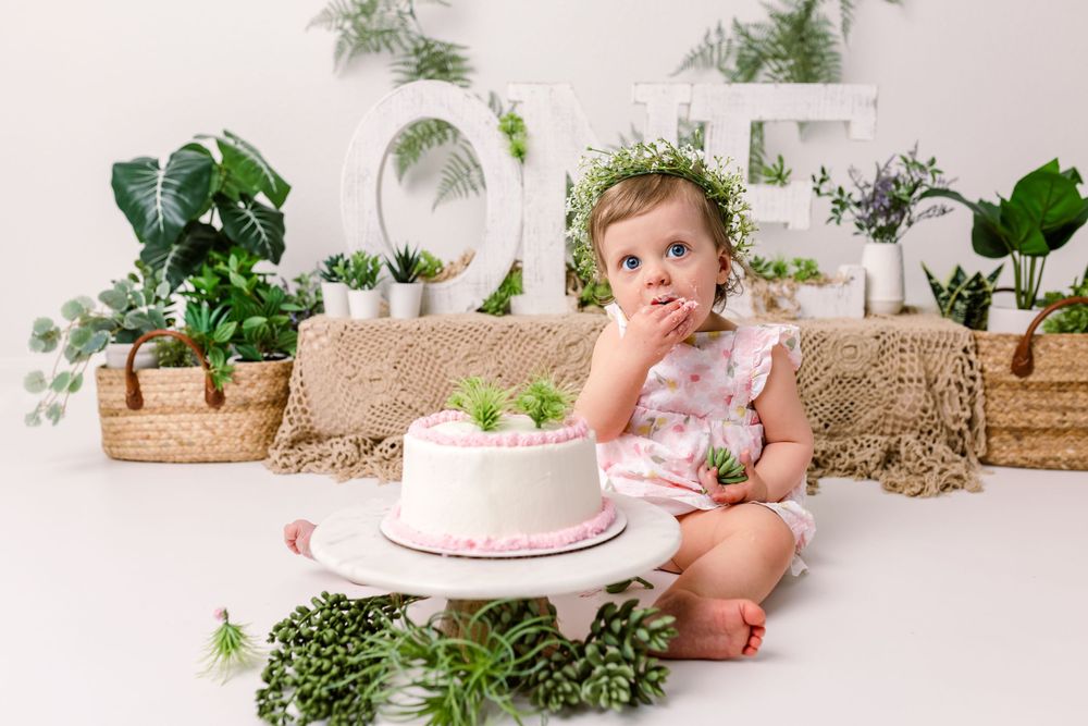 First birthday cake smash setup with pink cake against white backdrop decorated with potted plants and natural elements in cedar park tx
