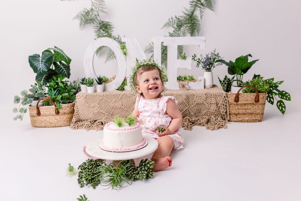 First birthday cake smash scene with ONE letters, potted plants and greenery on white backdrop with excited baby in cedar park tx studio