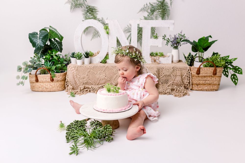 Cake smash photoshoot setup with white 'ONE' letters, potted greenery and a pink frosted birthday cake on white backdrop.