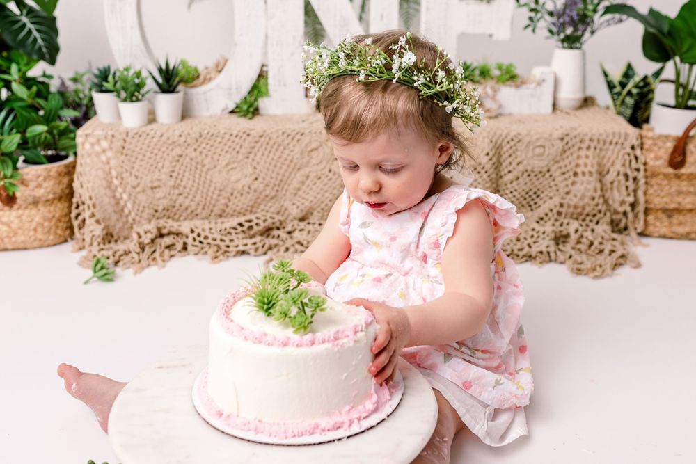 A delicate pink floral cake sits before a young girl wearing a pink dress and floral crown during her cake smash photos in cedar park tx