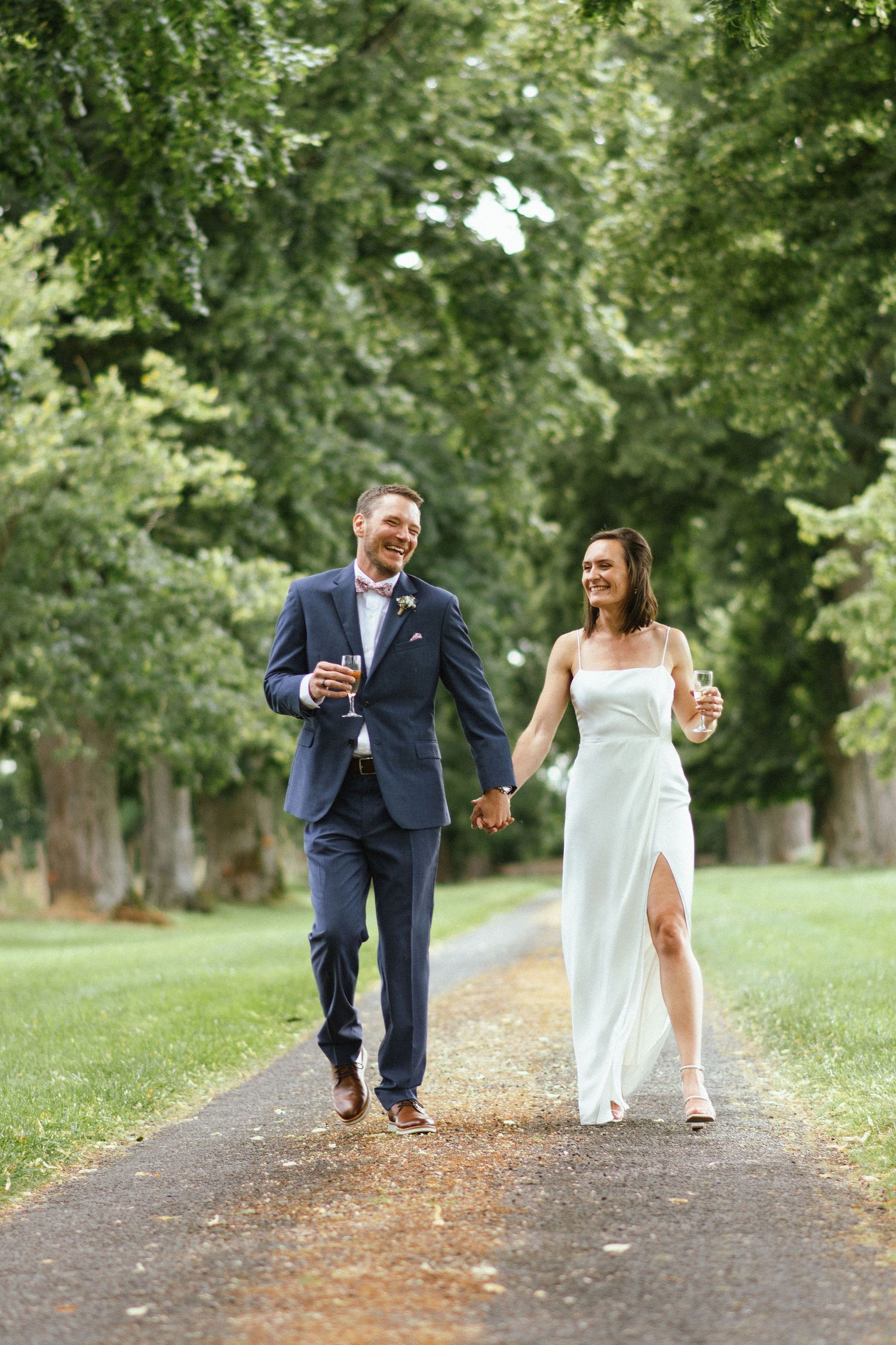 Couple de mariés marchant main dans la main sur une allée arborée, souriants et détendus.
