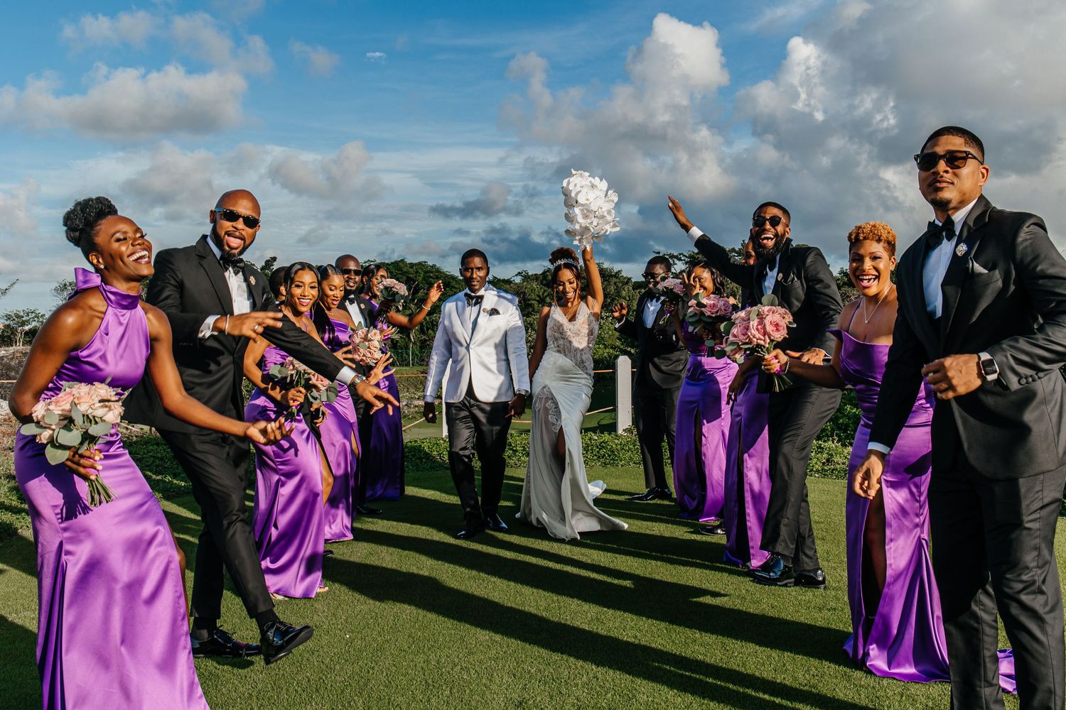 Wedding party in purple dresses celebrates on a golf course under blue skies.