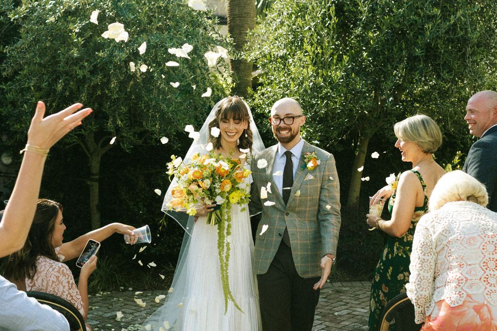 A bride and groom smiling ear to ear as they walk down the aisle of their wedding ceremony, while flower petals are thrown on them by guests