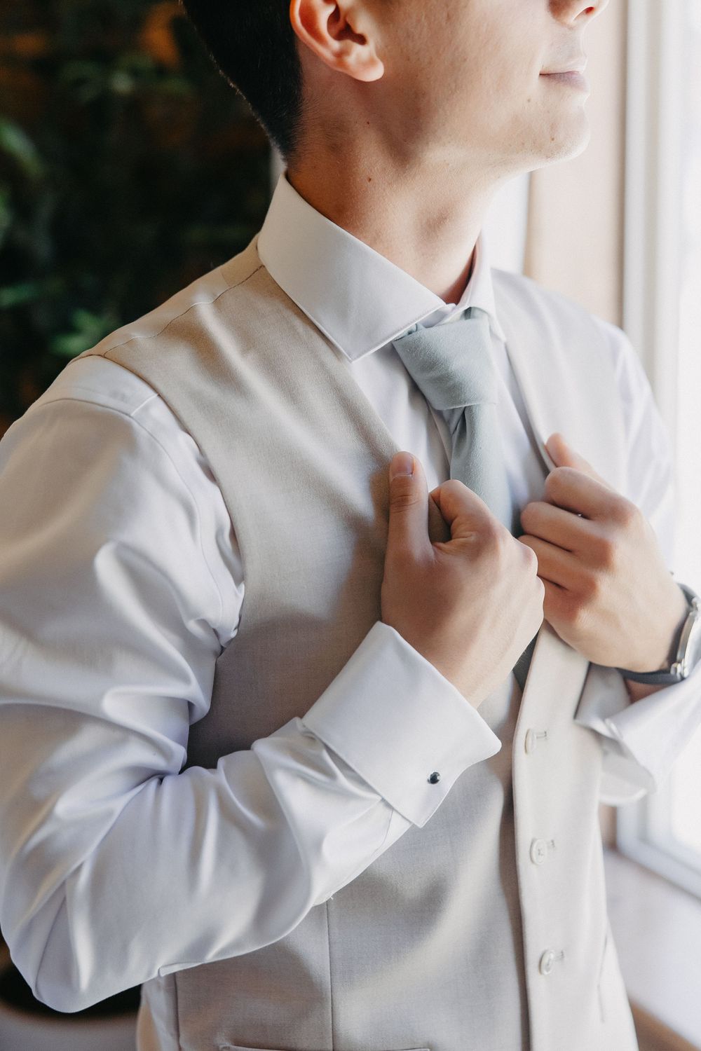 Close-up of hands adjusting a light blue tie on a crisp white dress shirt near a window.