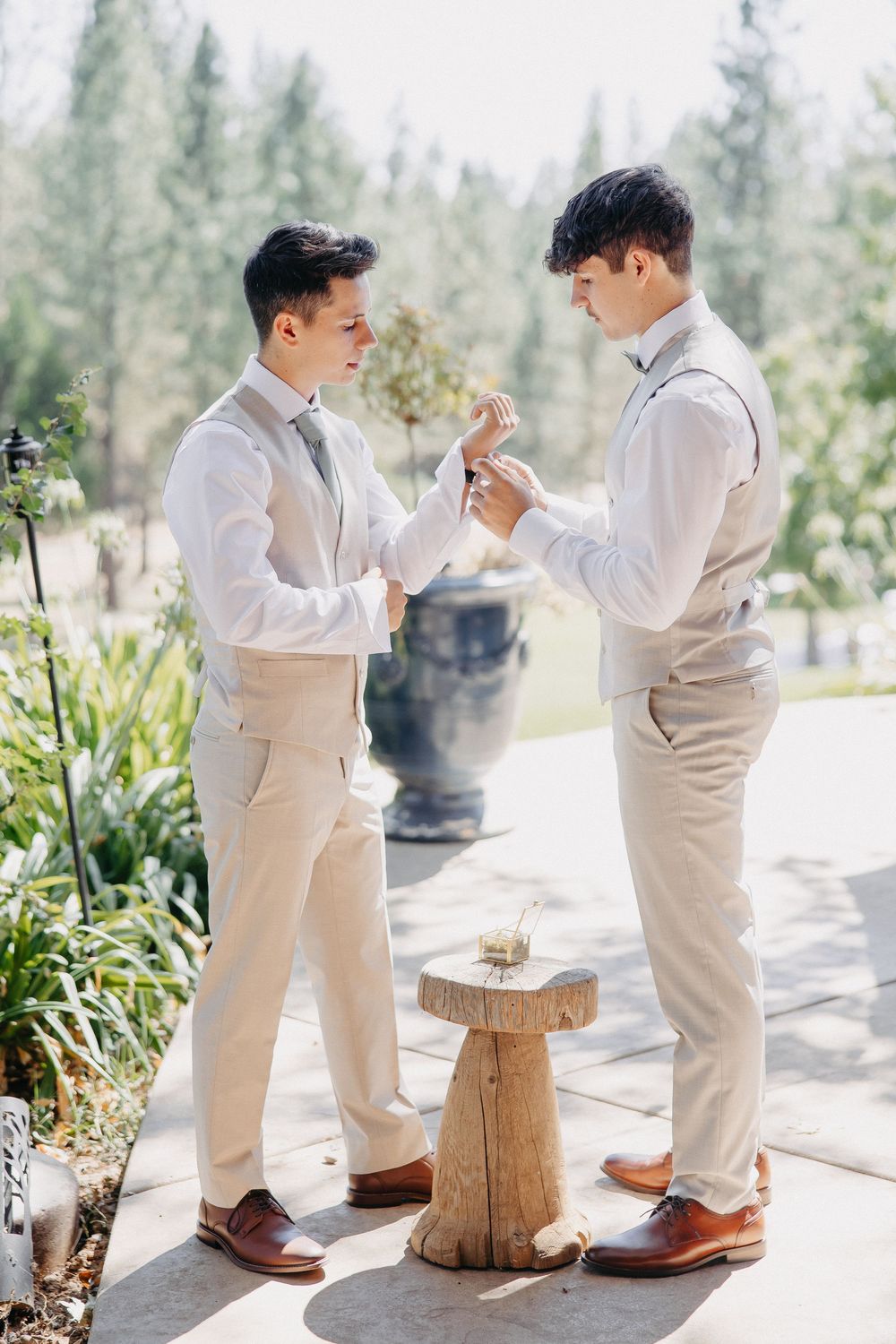 Two people in beige suits hold hands in a romantic moment during their wedding ceremony on a stone patio.