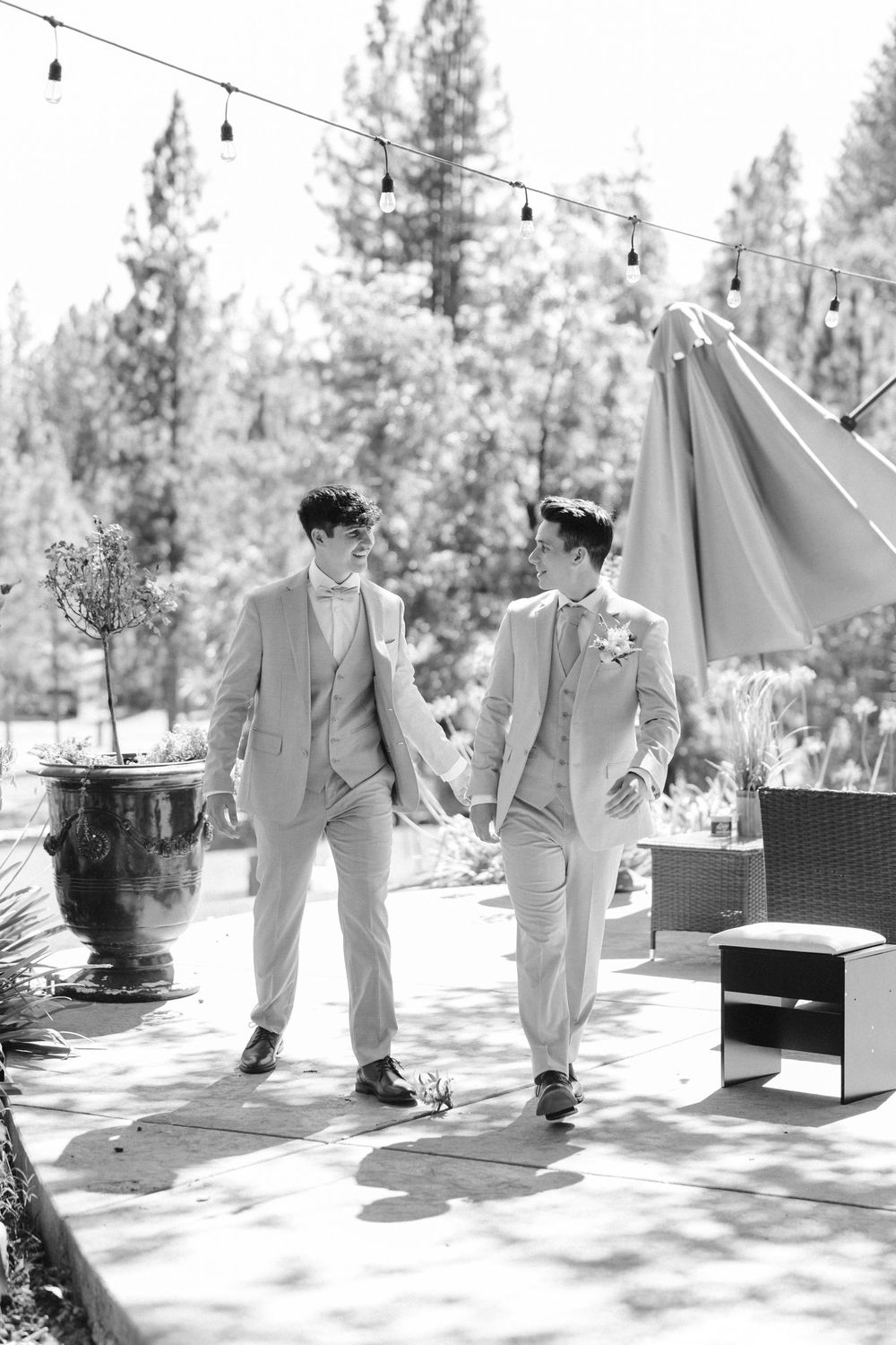 Two people in matching light gray suits walk together under string lights at an outdoor woodland venue.