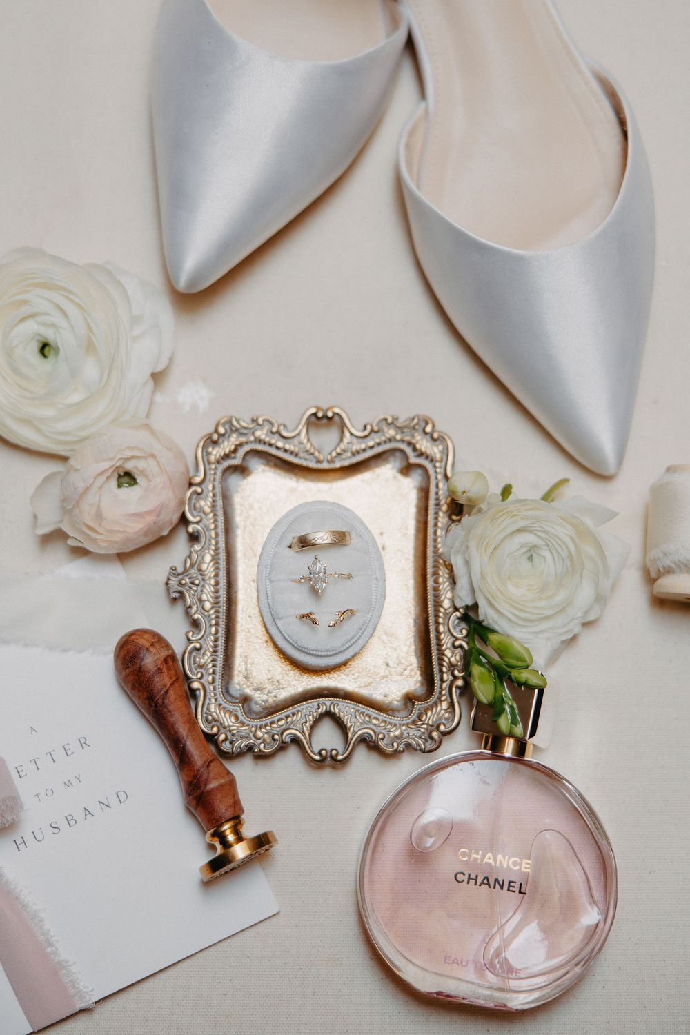 Wedding accessories including silver shoes, white flowers, ornate frame, and Chanel Chance perfume arranged on light surface.