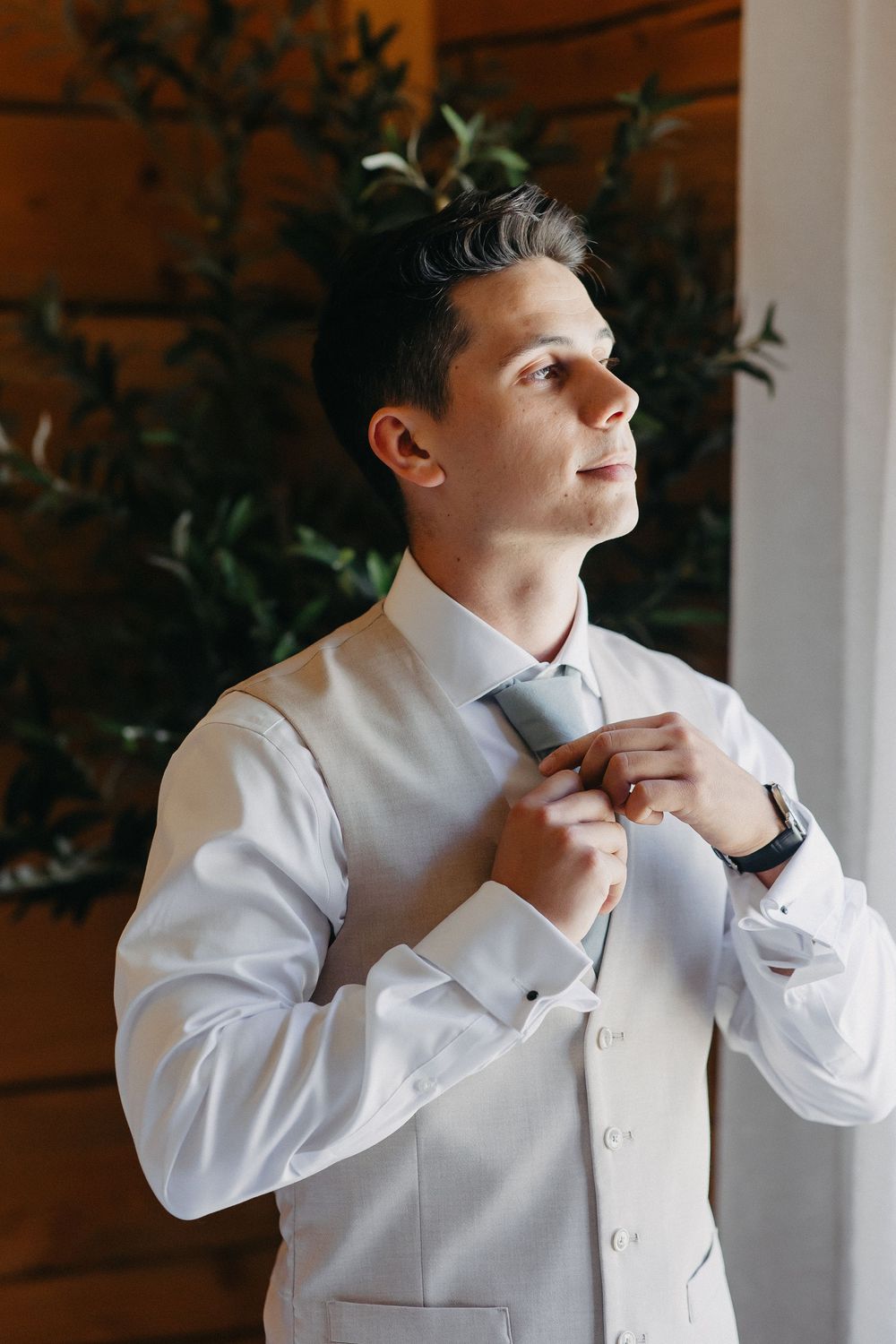 A groom adjusts his tie while getting ready in a room with natural lighting and indoor plants.