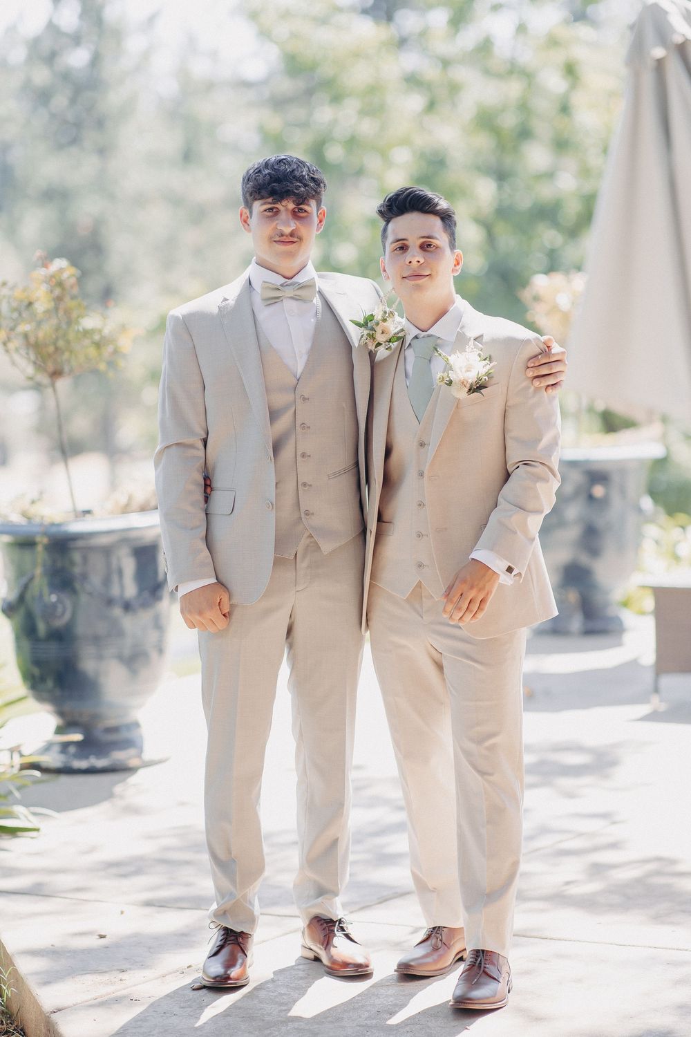 Two people wearing matching beige suits and bowties standing together on a sunny patio.