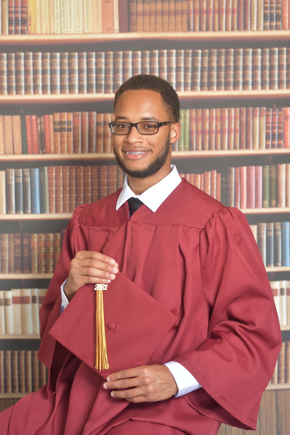 Graduate in burgundy cap and gown poses with diploma in library setting with bookshelves in background.