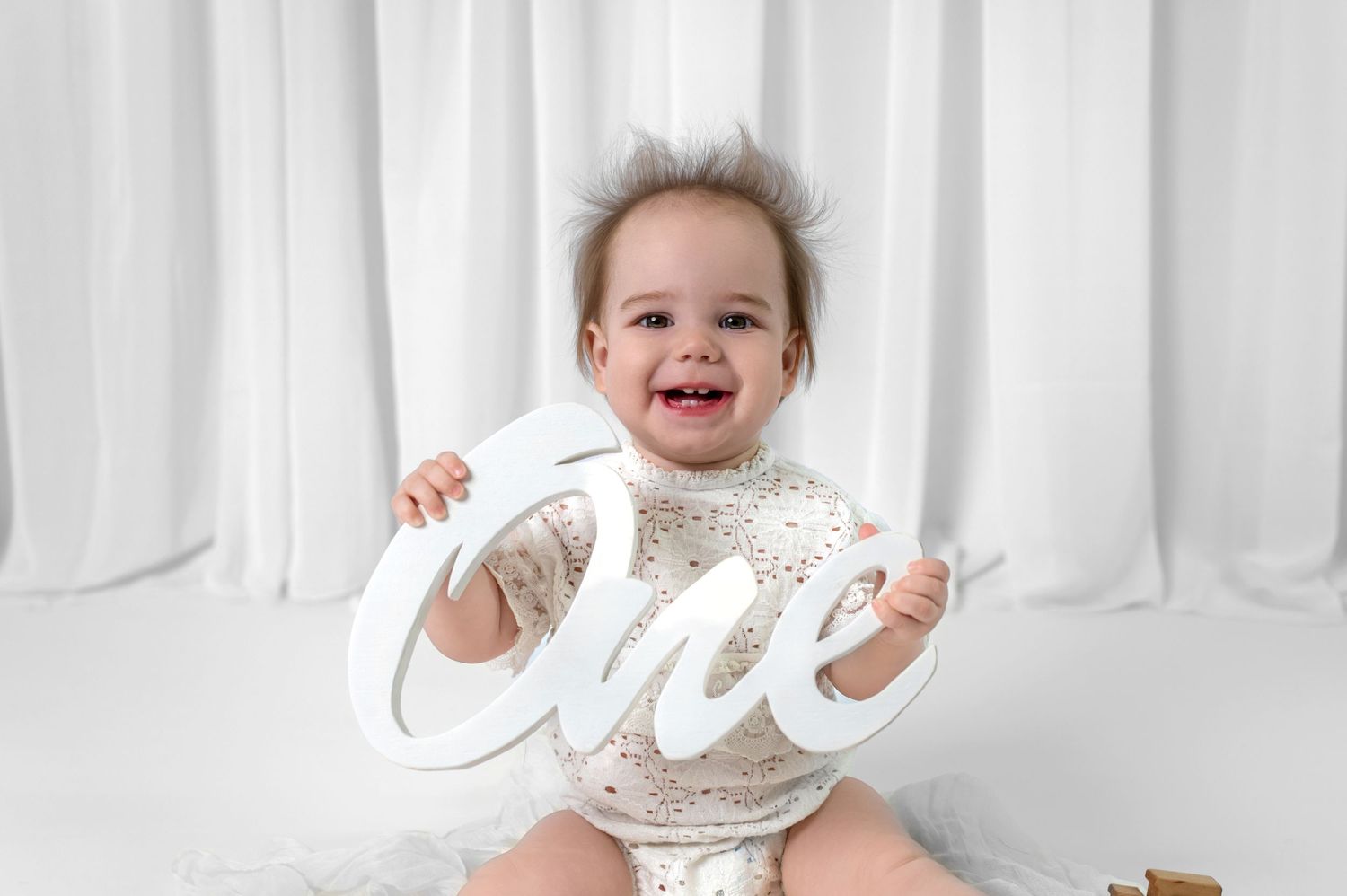 Happy baby in white outfit sits with 'One' sign against white backdrop during first birthday photoshoot.