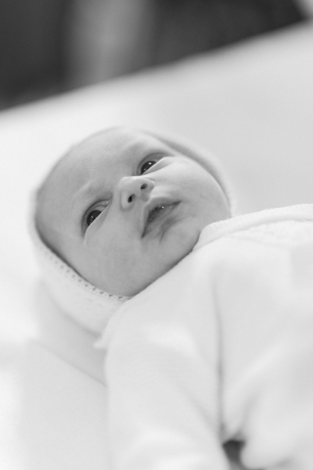 Close up black and white portrait of an alert newborn baby in white clothing.