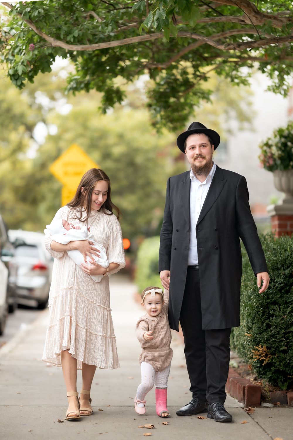 Family walking together on sidewalk with leafy trees overhead.