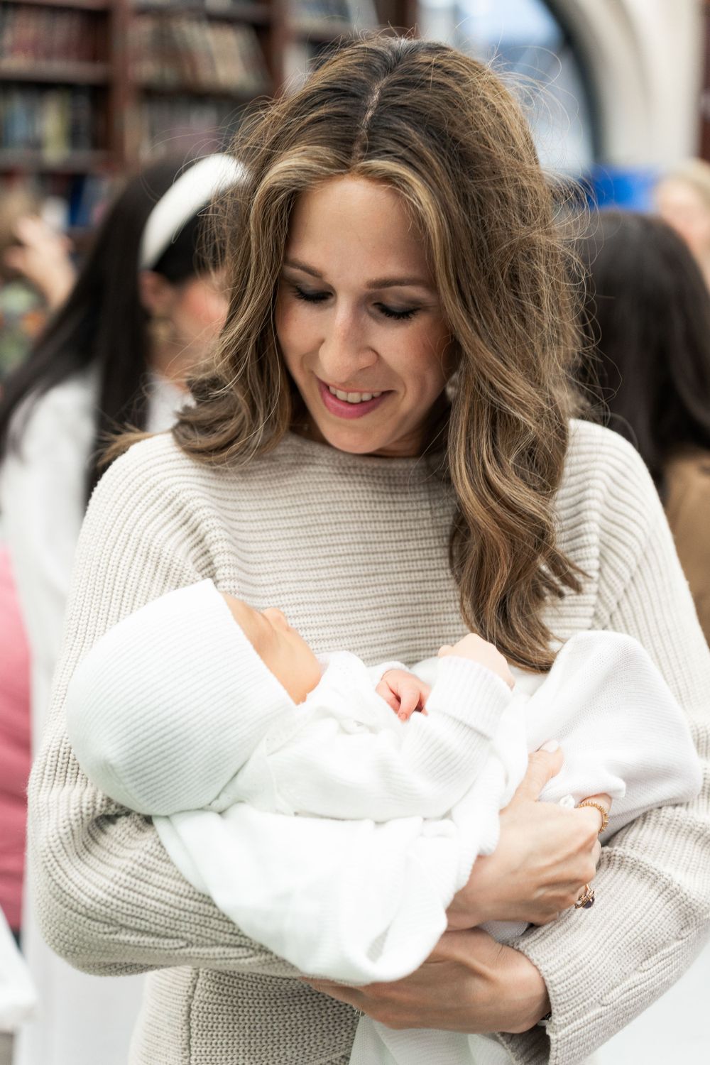 A mother in a beige sweater lovingly holds and smiles at a newborn baby wrapped in white blankets.