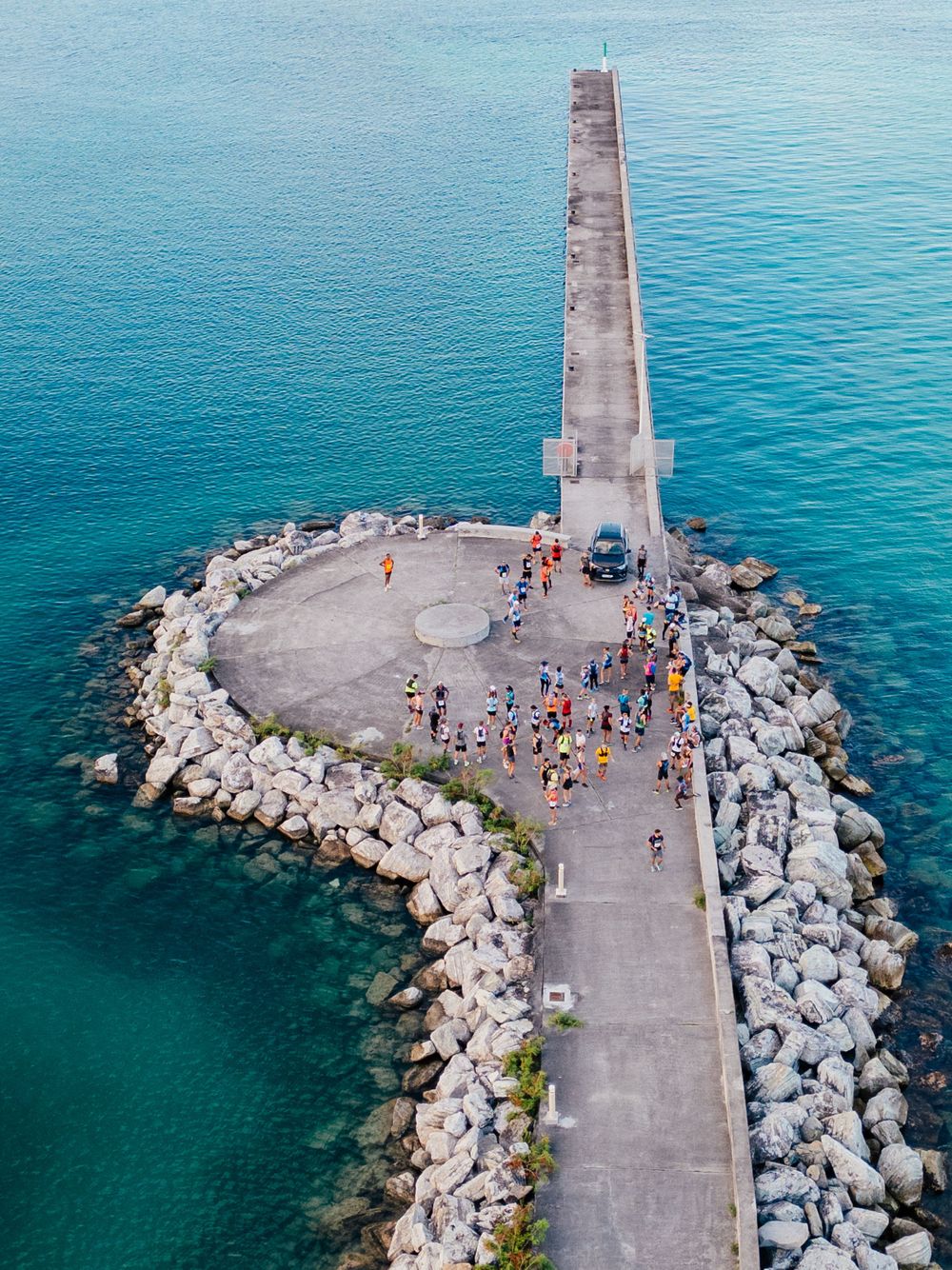 Aerial view of people gathered on a concrete pier with breakwater rocks surrounded by turquoise ocean waters.
