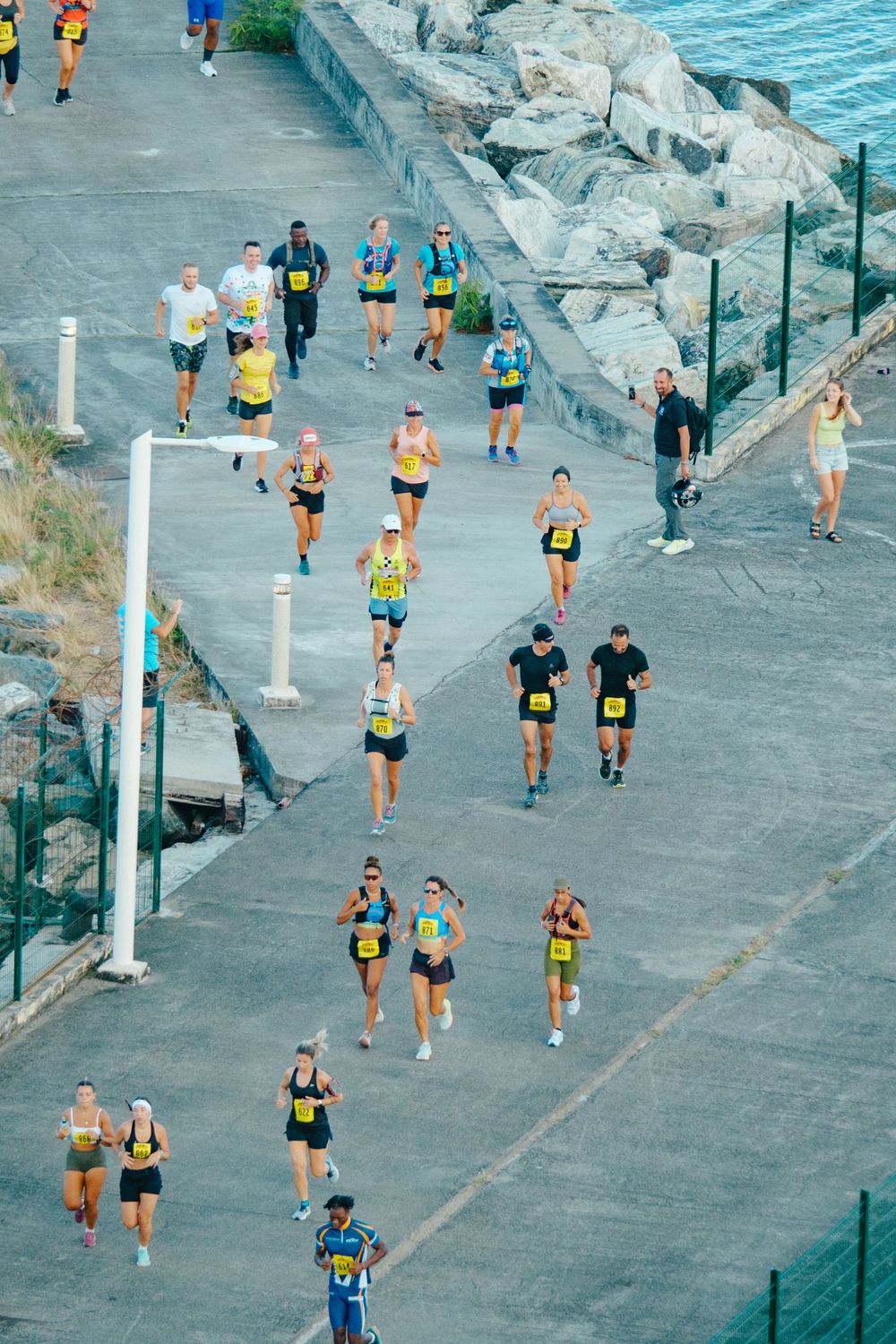 Runners compete in a coastal road race along a path next to rocky shores and turquoise waters.