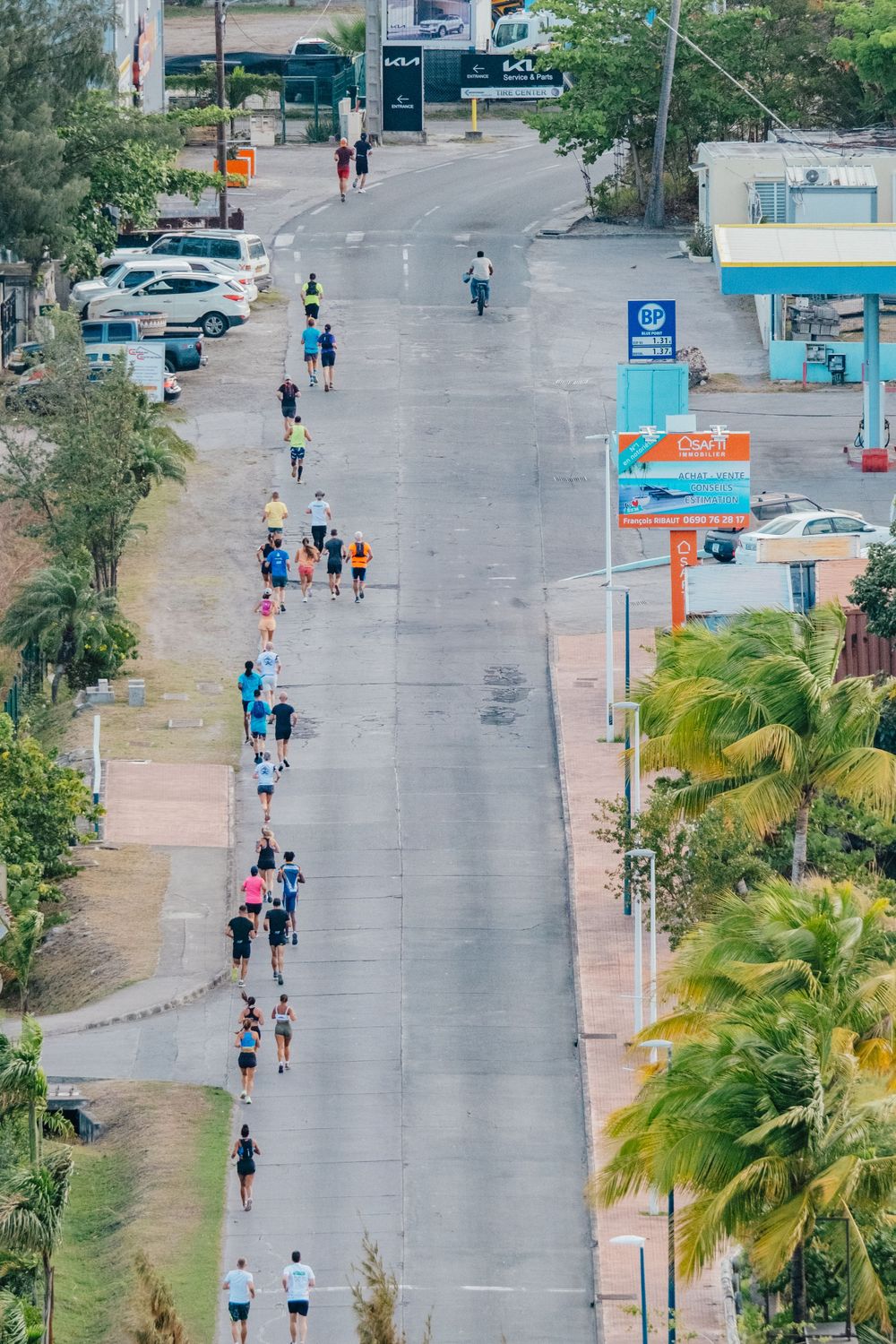 Aerial view of people jogging and walking along a palm-lined street with parked cars and buildings visible.