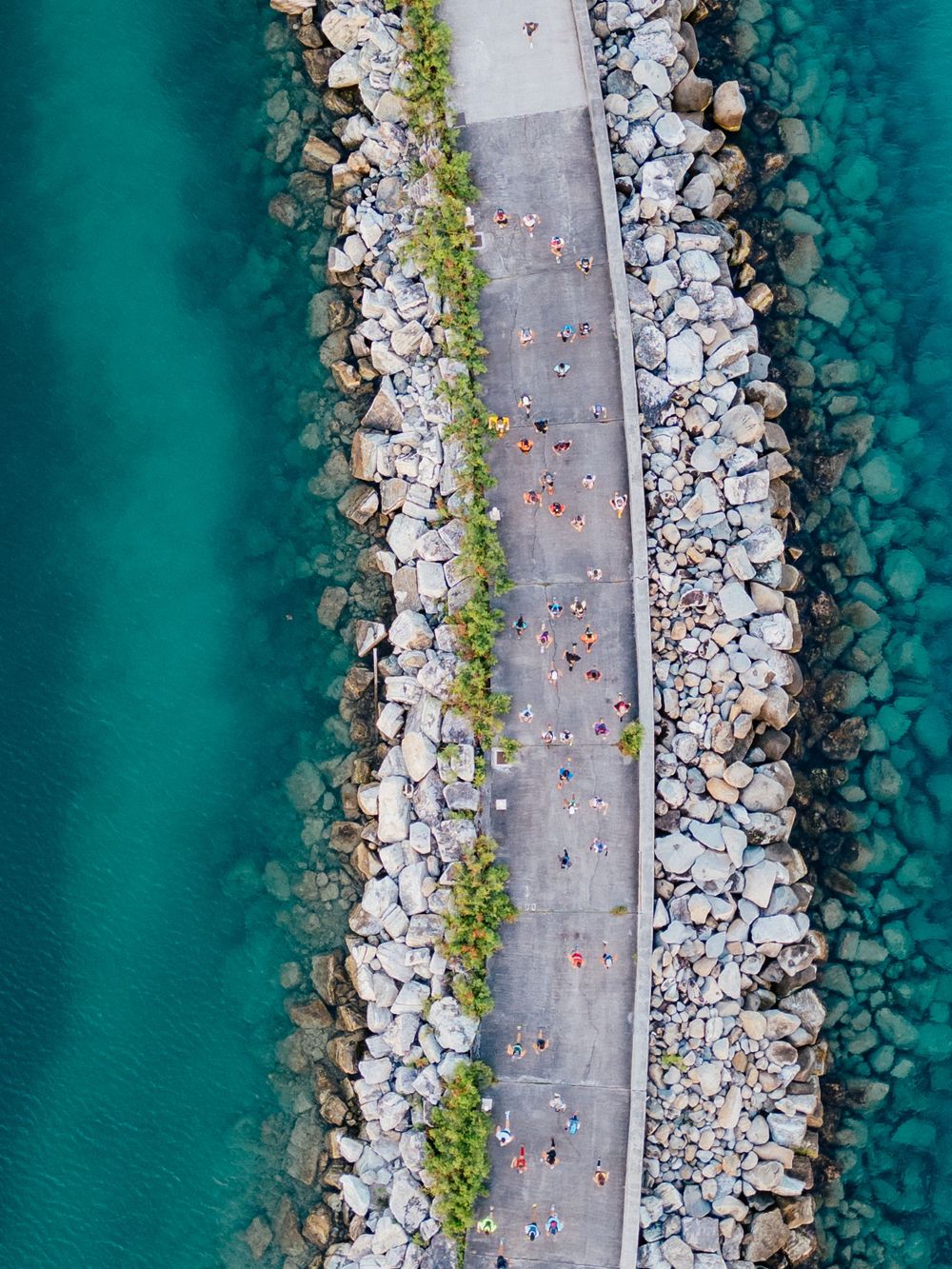 Aerial view of a stone pier walkway between turquoise waters with small figures walking along the path.