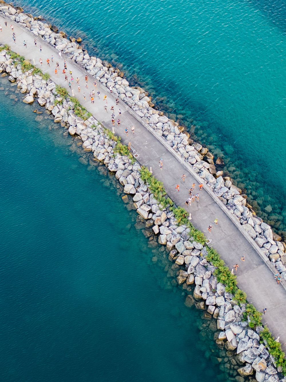 Aerial view of curved stone breakwater with walking path along turquoise Mediterranean waters.