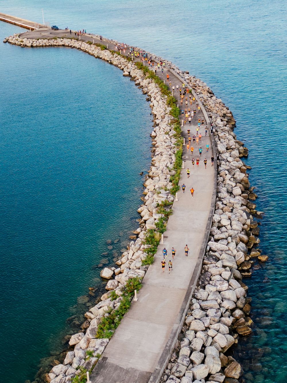 Aerial view of a curved concrete breakwater path lined with rocks and dotted with pedestrians along blue ocean waters.