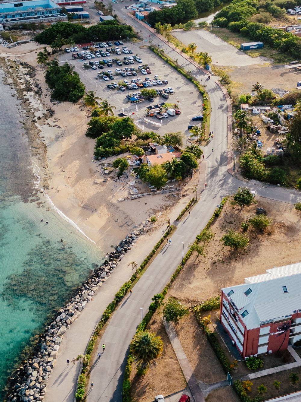 Aerial view of a coastal road with parking lot, turquoise waters, and rocky shoreline in a tropical setting.