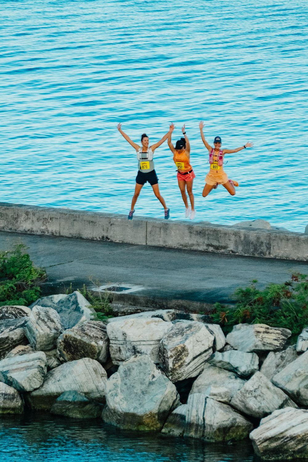 Three friends leap joyfully from a concrete wall near turquoise waters and rocky shoreline on a bright summer day.