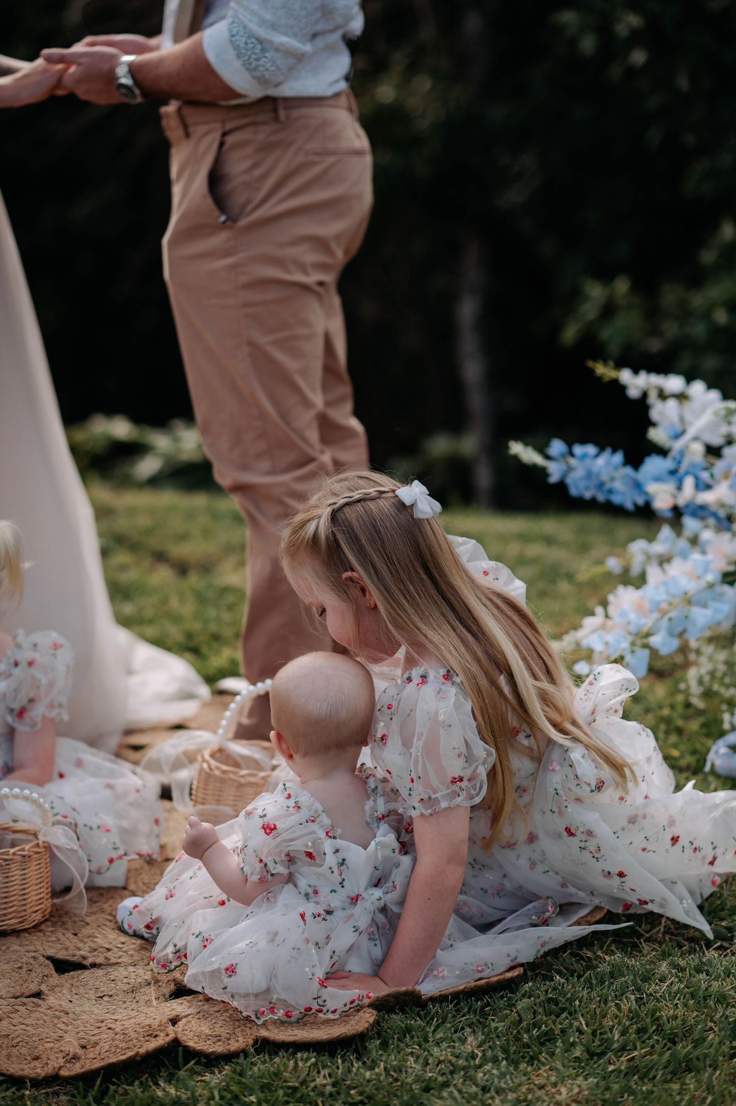 Children in floral dresses play on a picnic blanket during an outdoor wedding.
