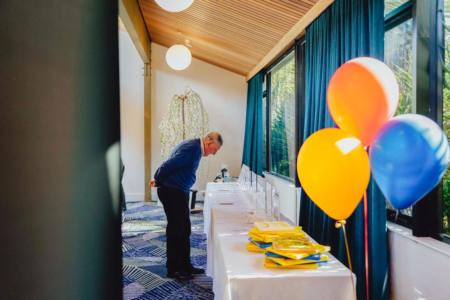 A person setting up a party table with colorful balloons and decorations in a room with blue curtains.