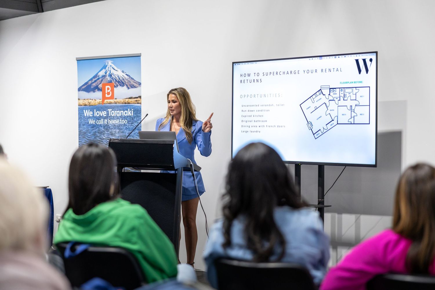 A presenter speaks to an audience while standing near digital displays during an educational presentation.