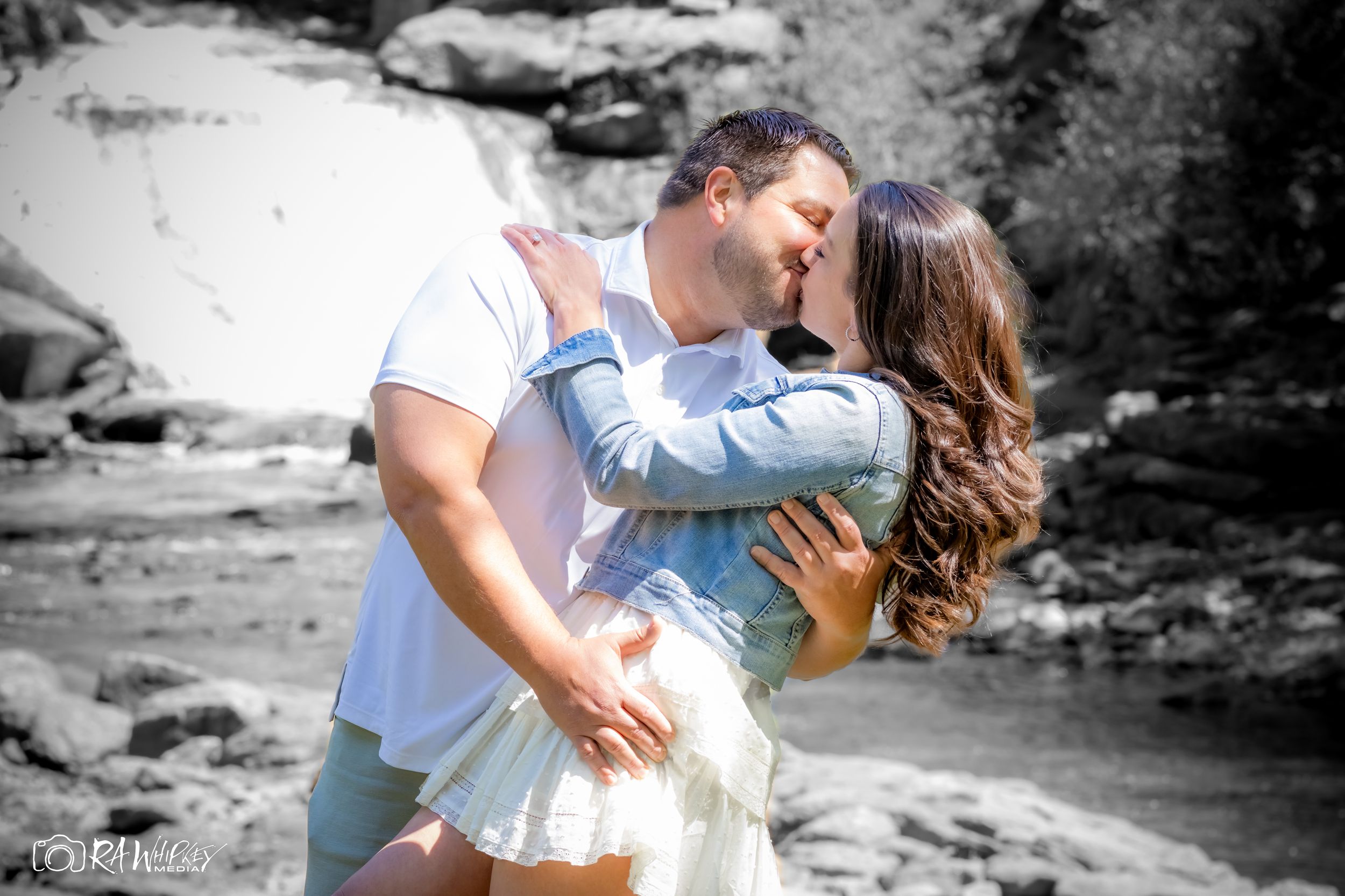 A romantic black and white photo of a couple embracing near rocky waterfall scenery.