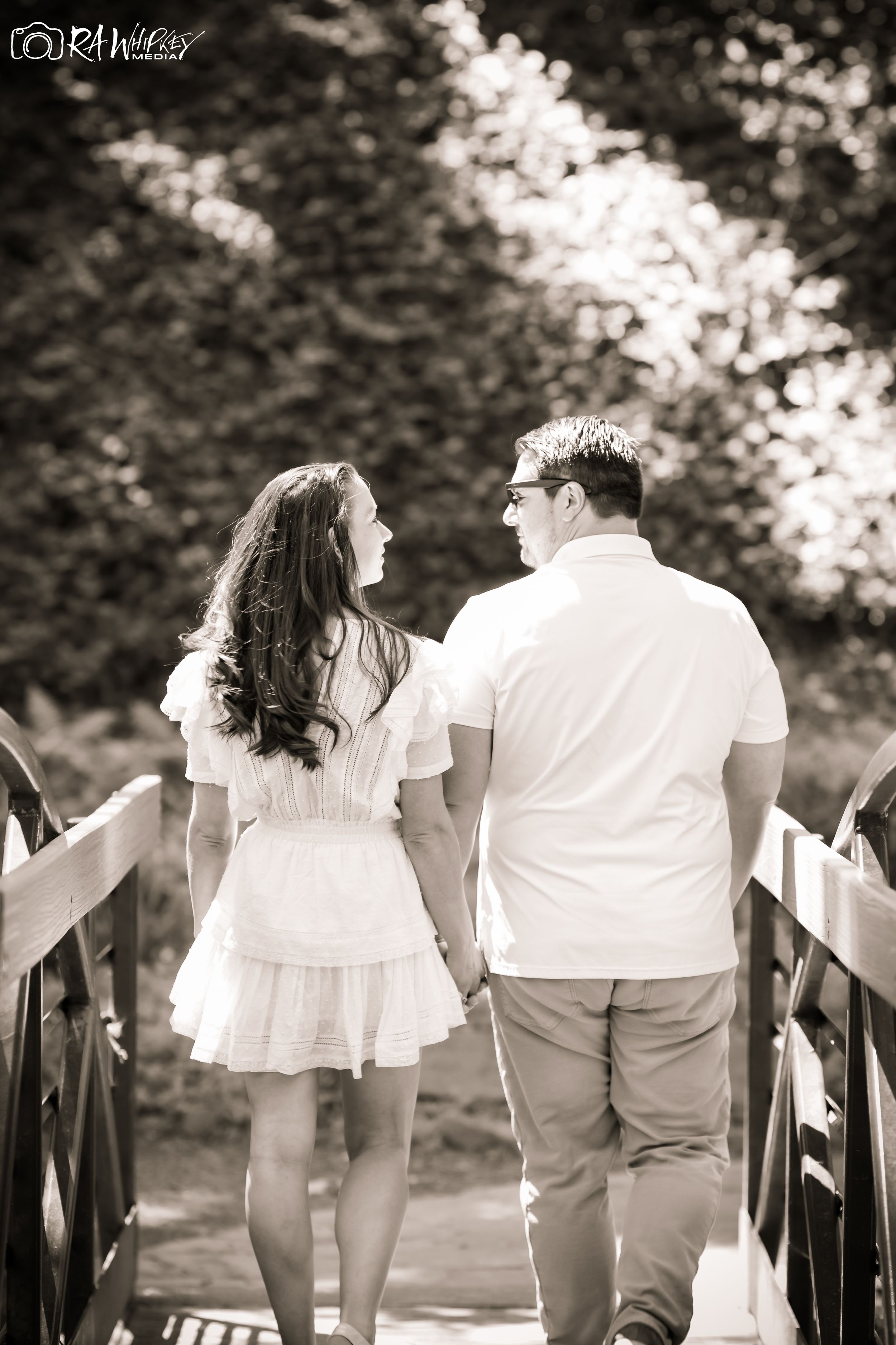 A black and white photo of a couple walking together on a wooden bridge in casual attire.