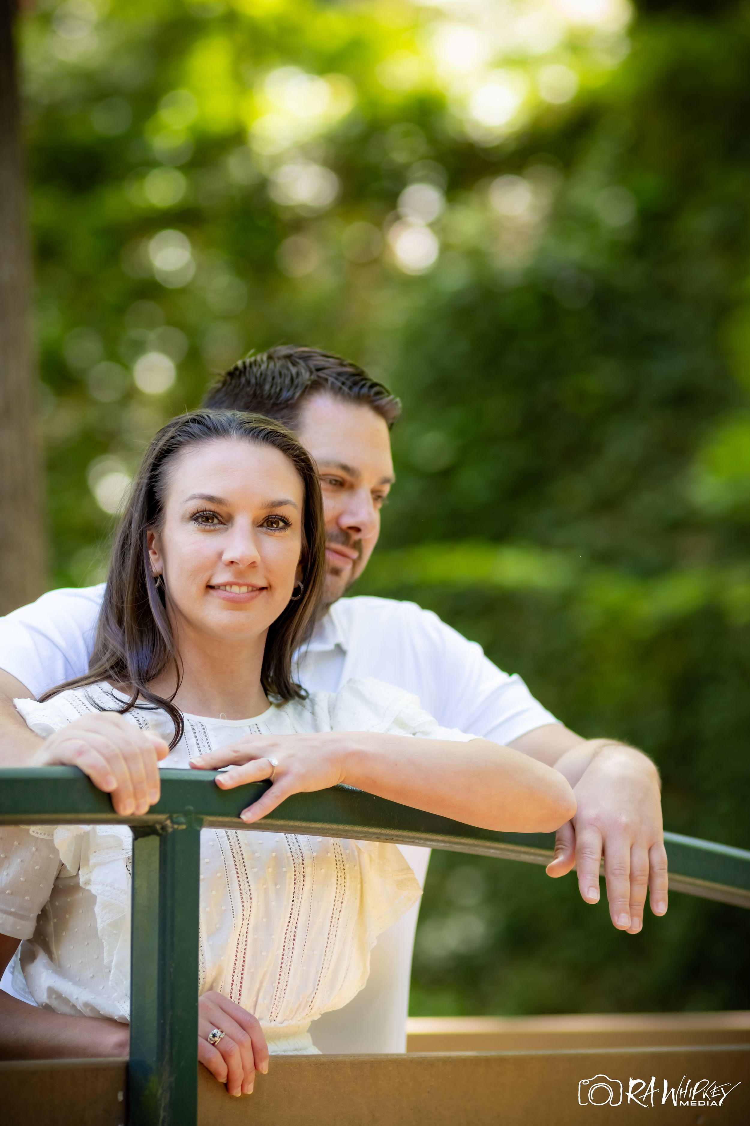 A tender moment between a couple leaning on a bridge railing surrounded by lush greenery.