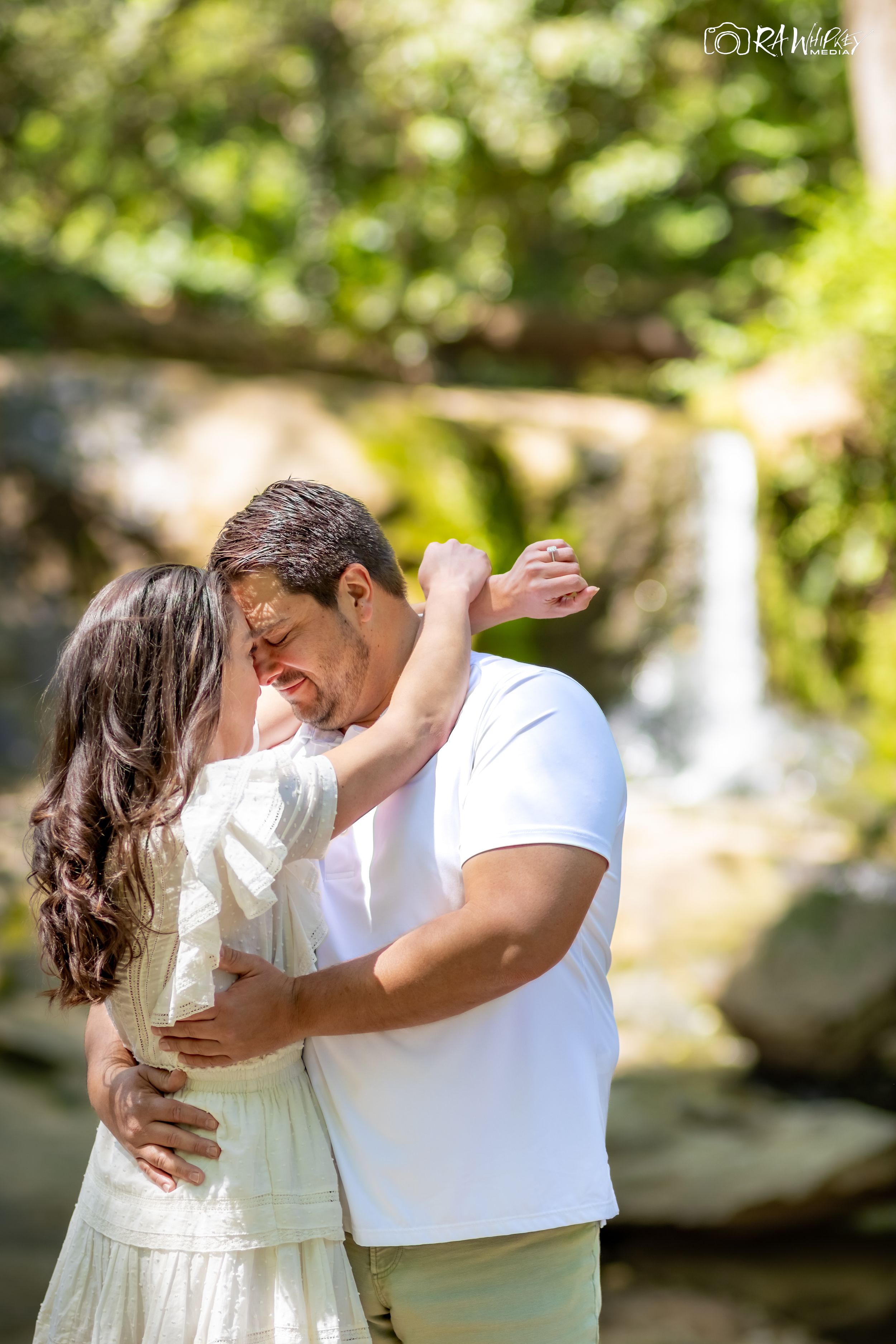 A romantic embrace between a couple in white outfits with a waterfall in the background.