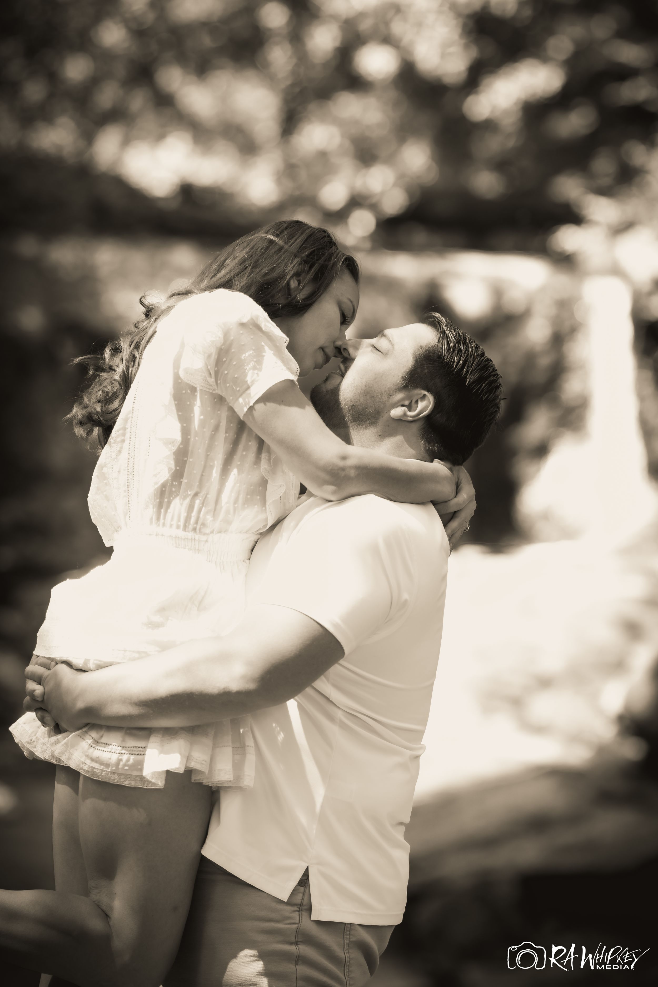 A romantic couple shares an intimate embrace in a soft black and white outdoor portrait.