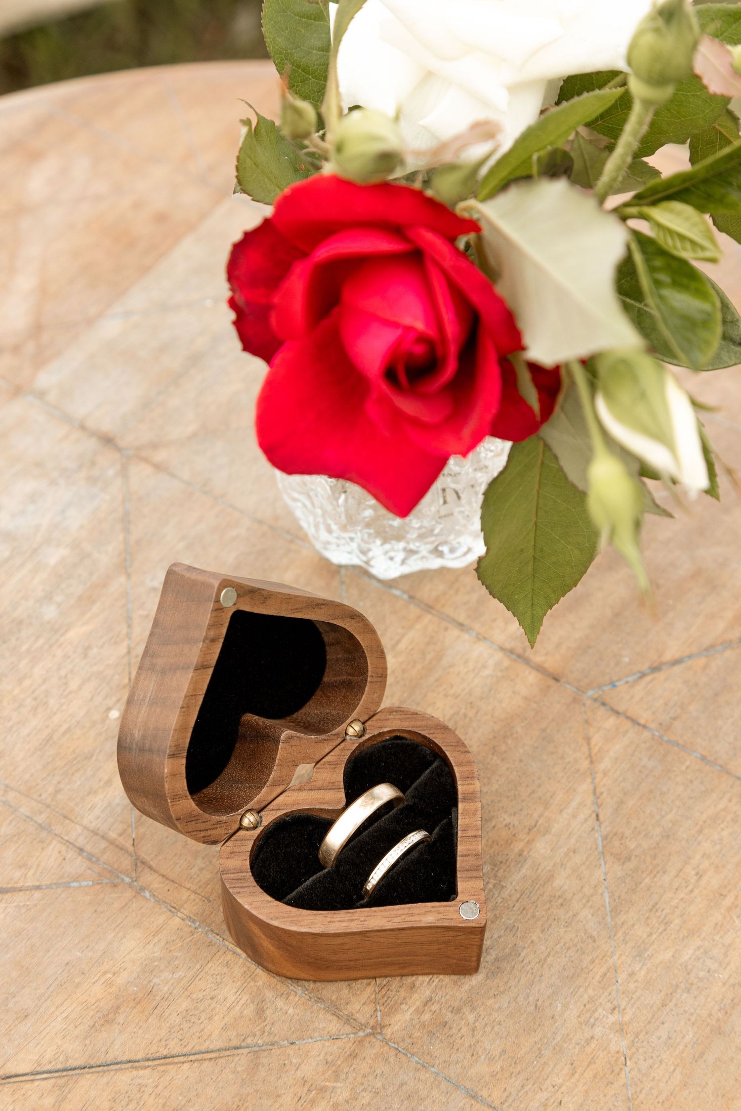 Wedding rings in heart-shaped wooden box next to red rose in lace-wrapped vase on wooden surface.