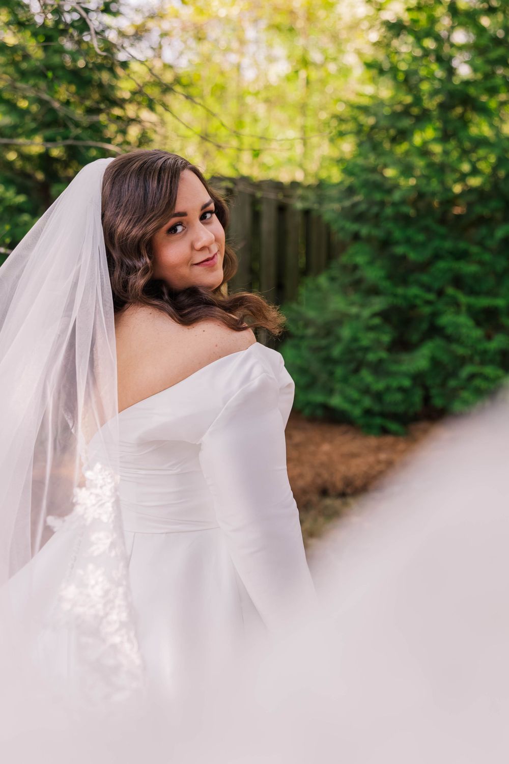 Bride in flowing white veil poses outdoors against green garden backdrop.