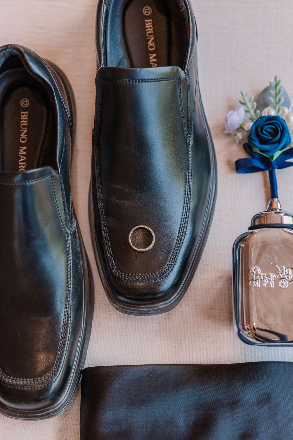Black formal dress shoes and a blue boutonniere arranged with a cologne bottle on light fabric.