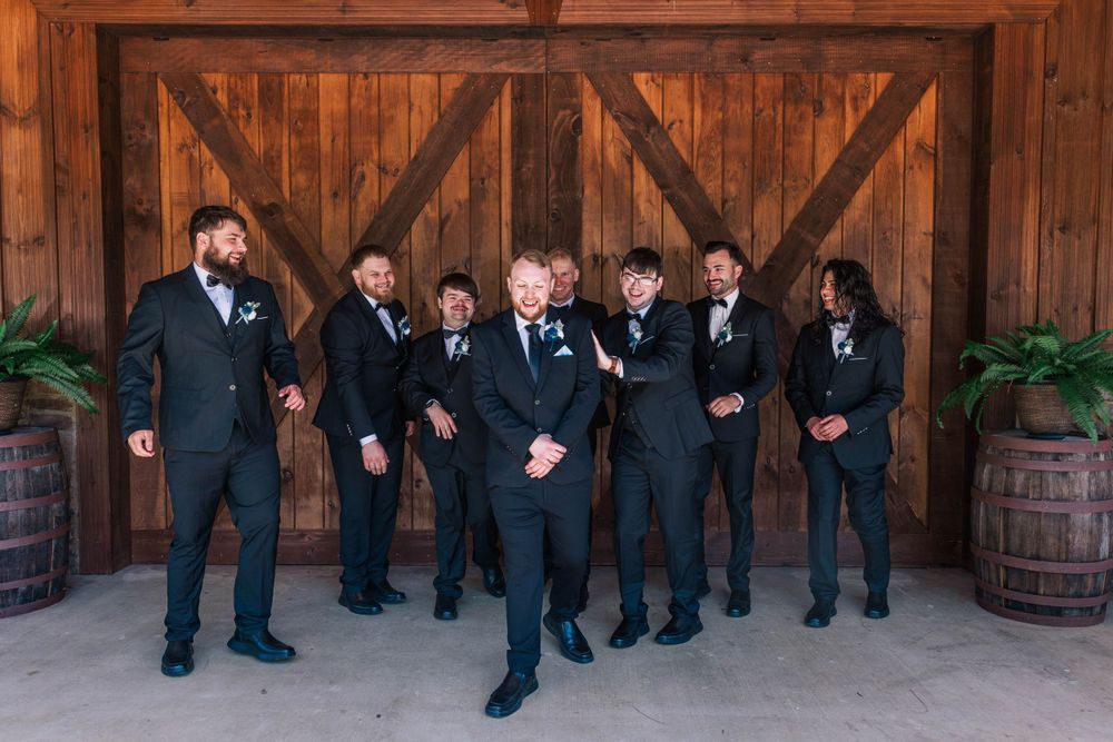 Groomsmen in black suits posing informally against a wooden barn backdrop.